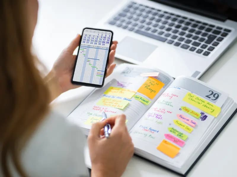 Scheduling software systems shown on a smartphone and planner as a woman organizes tasks with color-coded notes